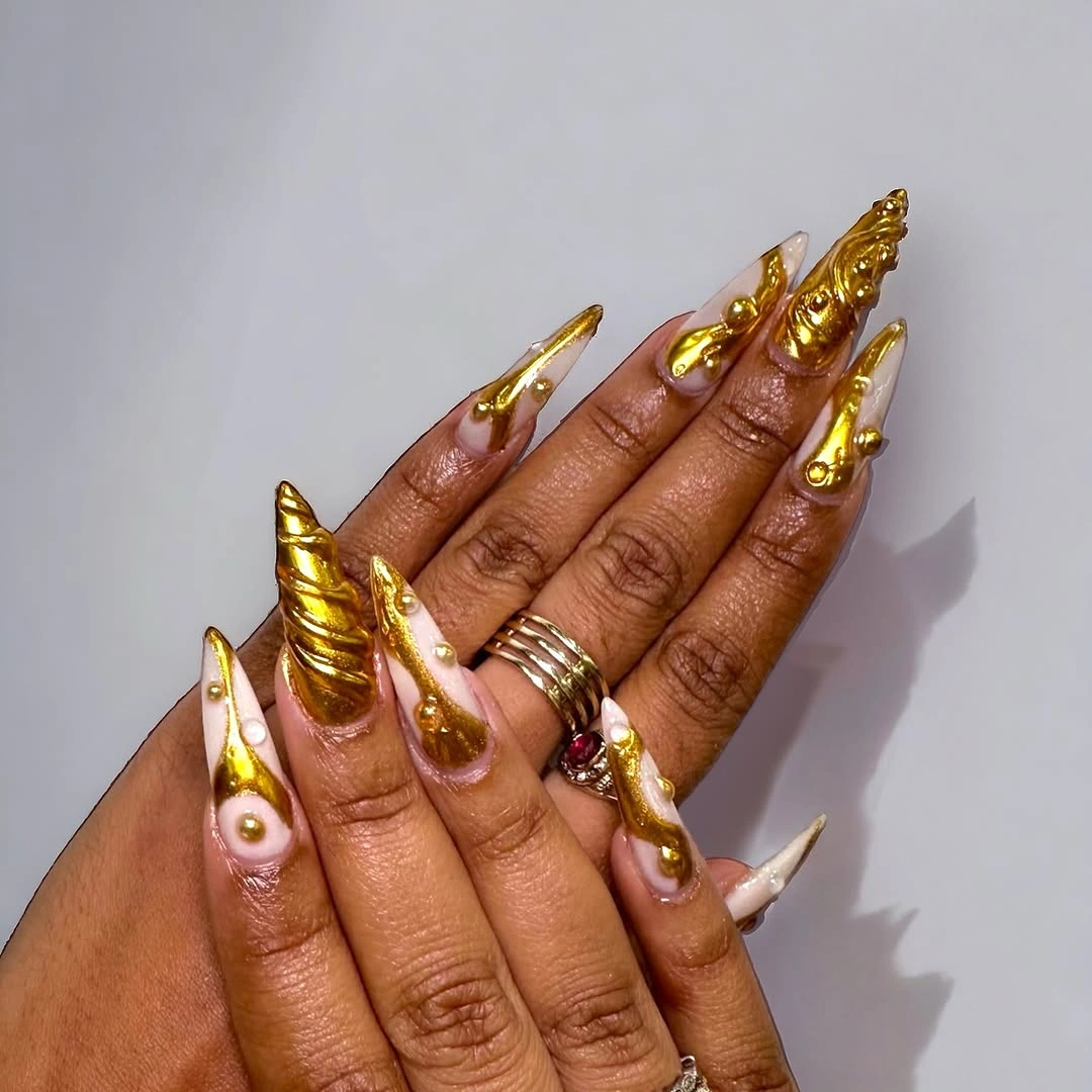 Extra-long pointed nude nails with sculptural gold-toned details and round accents