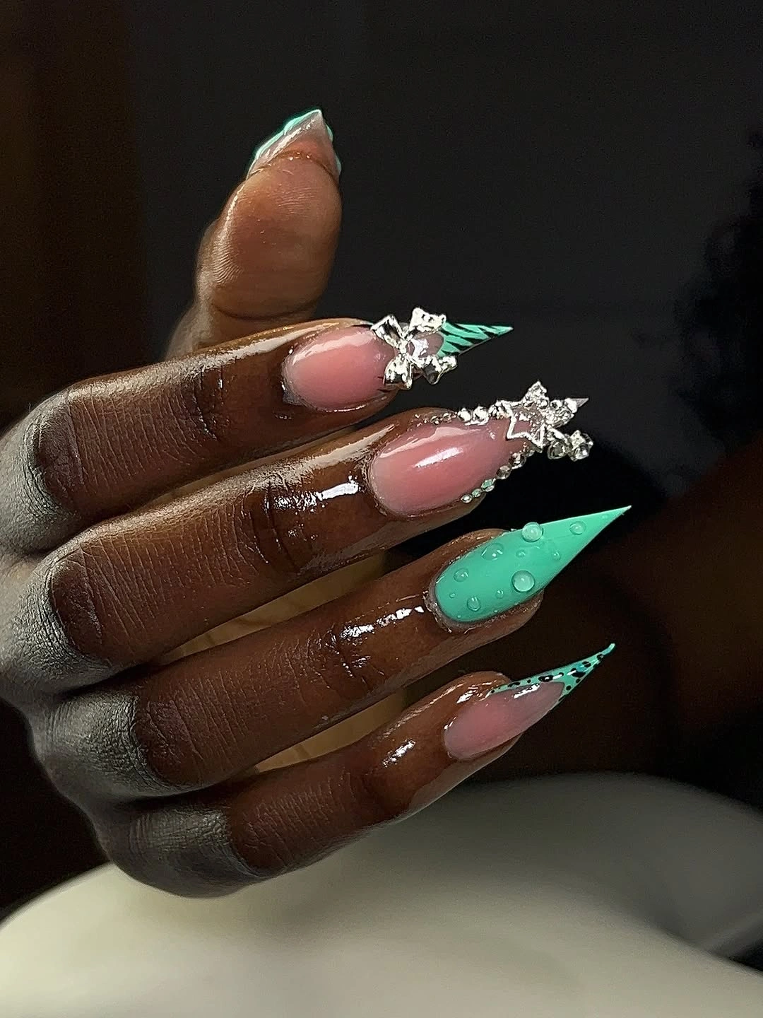 Long pointed nails alternating blush pink, emerald green, and small sparkling accents