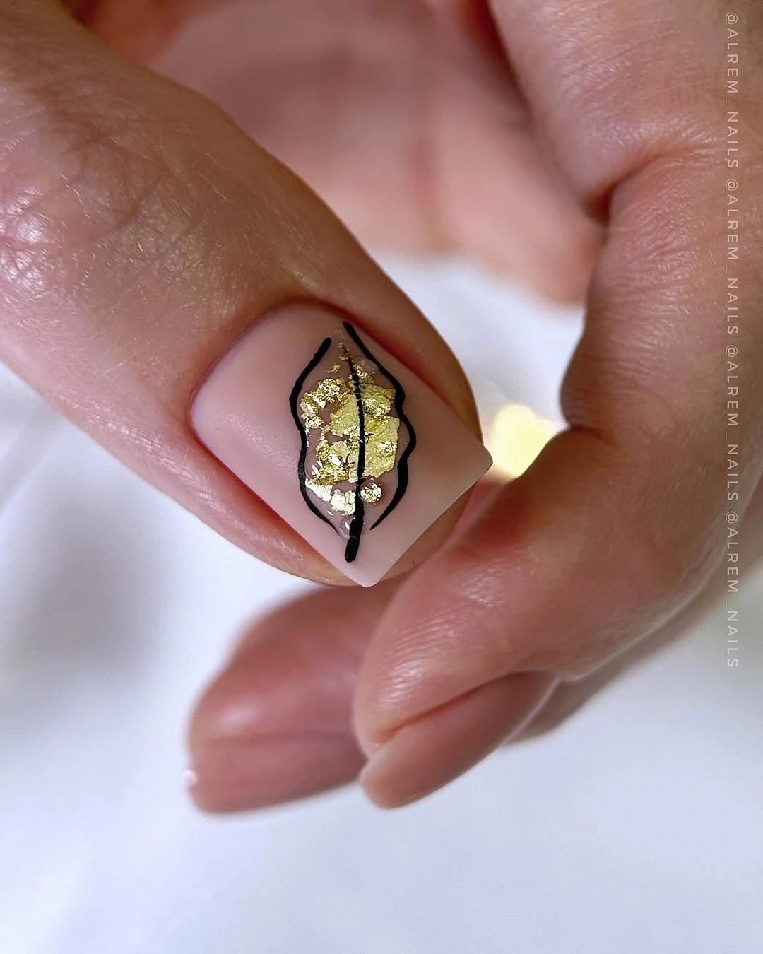 Pale nude nail with irregular black frame and small metallic gold leaf detail