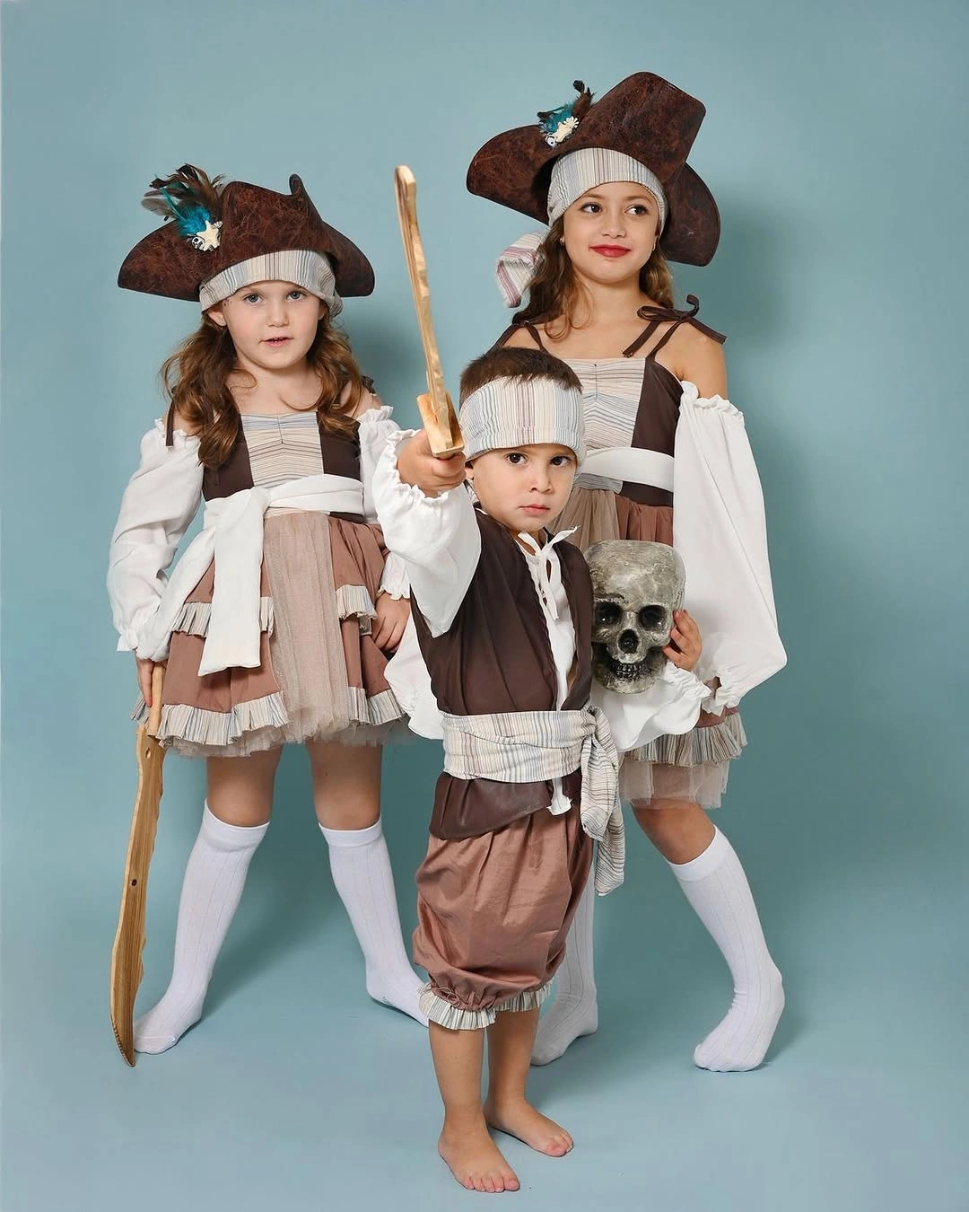 Three children in coordinated pirate costumes with head scarves, sword, and skull prop