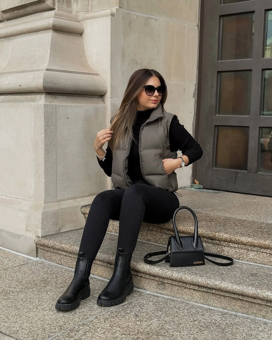 Olive-gray padded vest with black top, fitted pants, boots, and handbag