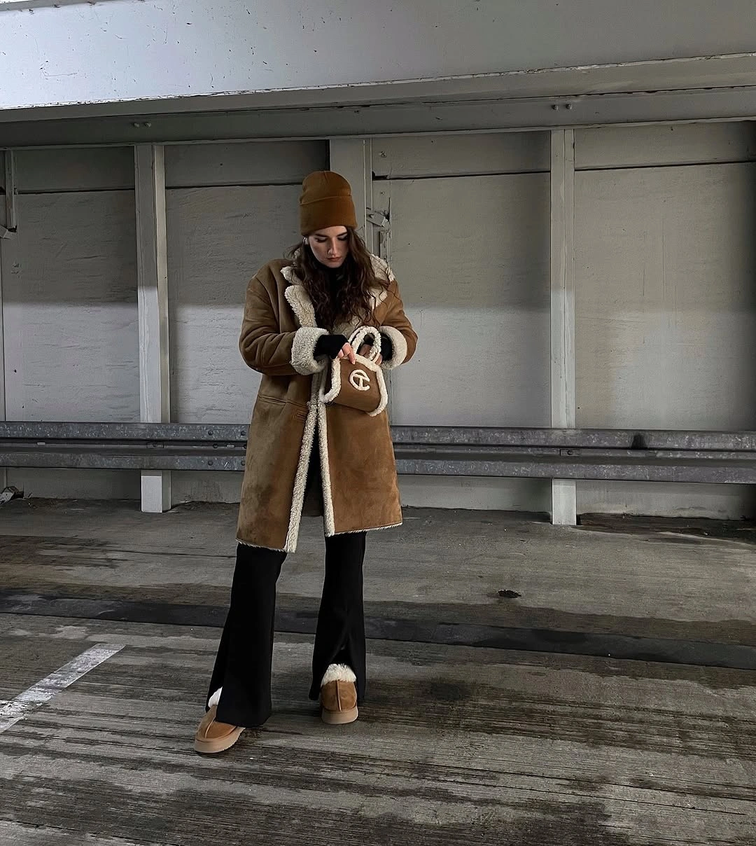 Brown cream-trimmed coat, mustard-brown beanie, flared black trousers, tan boots, and brown shoulder bag.