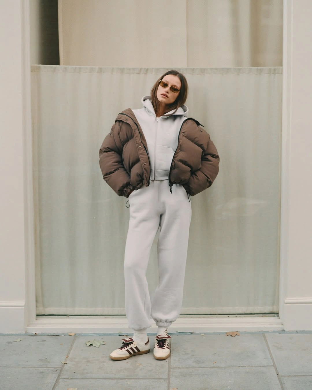 Cropped brown puffer over pale hoodie and joggers with white sneakers and dark sunglasses.