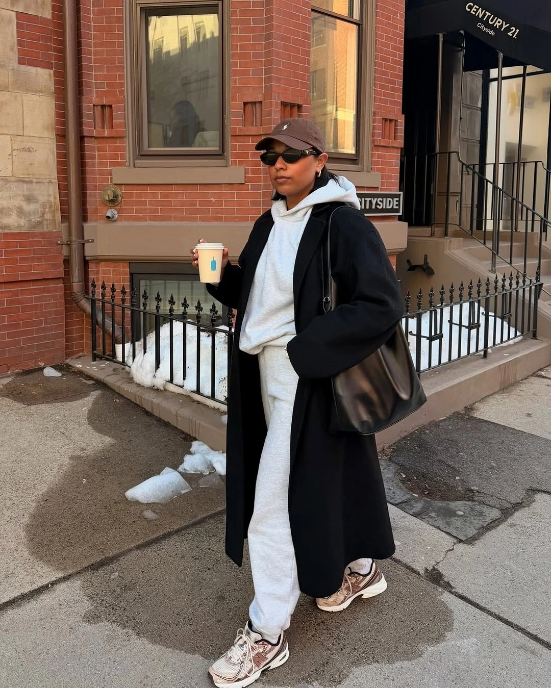 Long black coat, pale tracksuit, white sneakers, brown cap, black bag, and dark sunglasses.