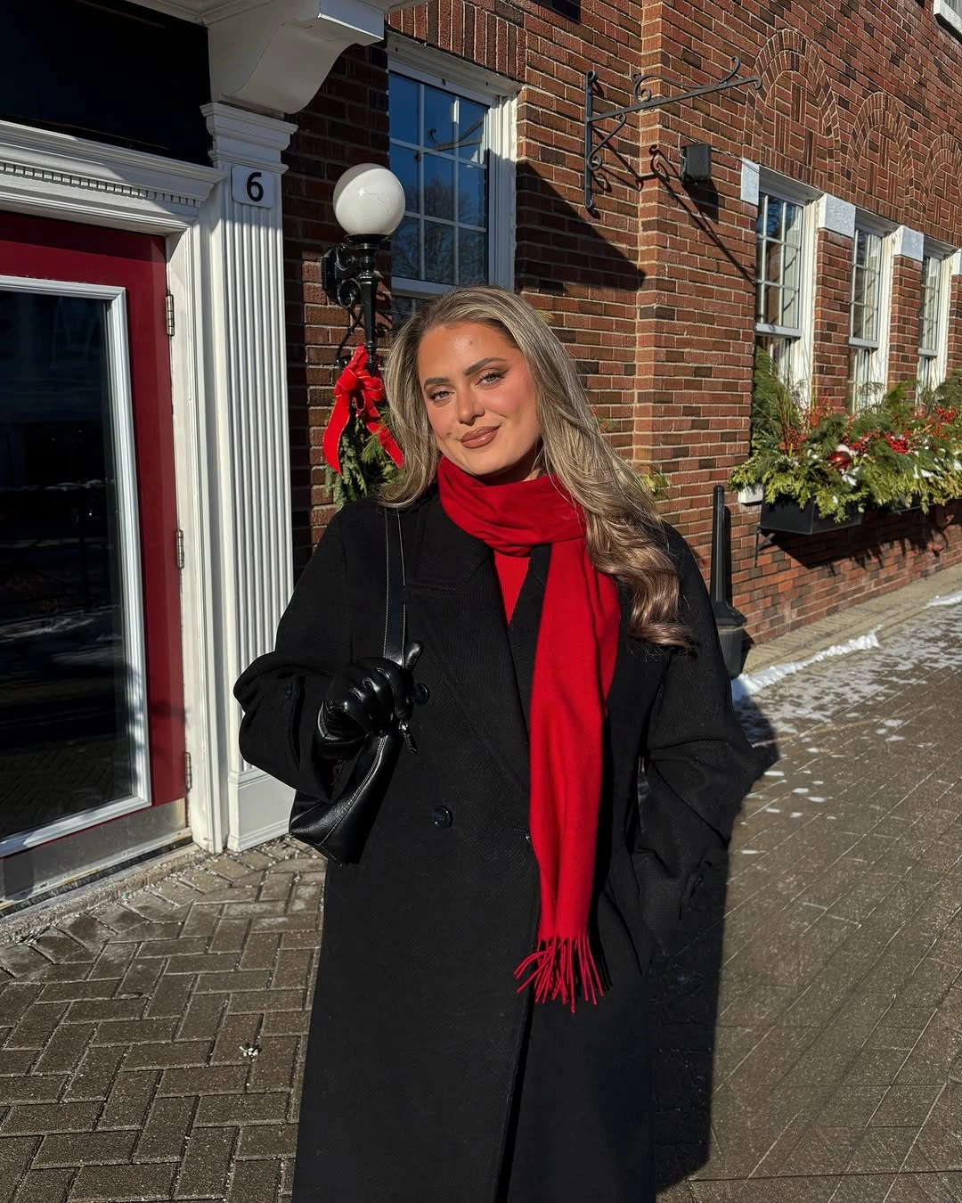 Long black coat with red fringed scarf, black gloves, shoulder bag, and dark sunglasses.