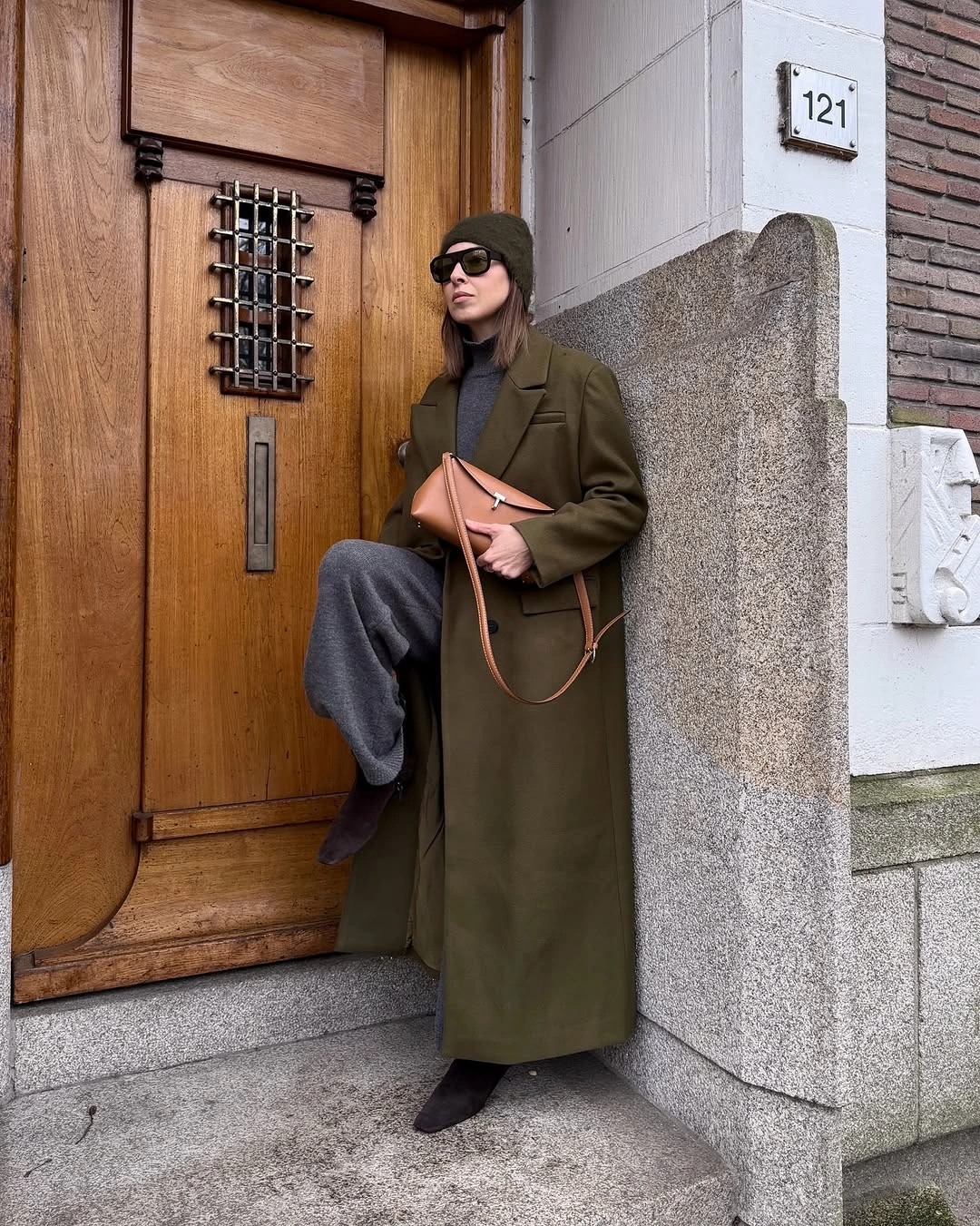 Olive long coat, gray trousers, beanie, and tan handbag