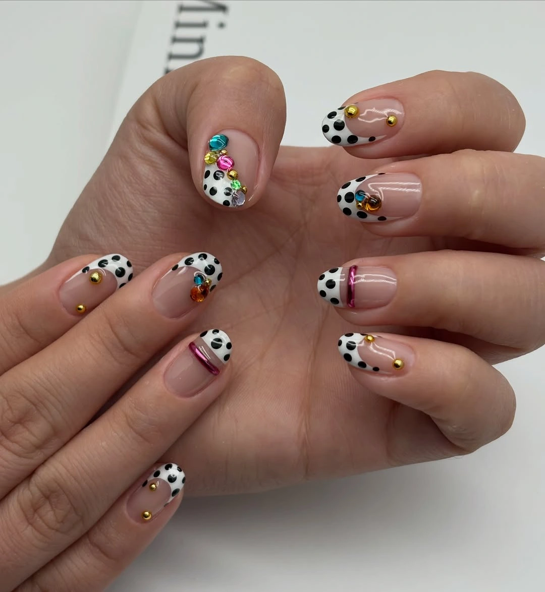 Short nude nails with black dotted white tips, small gold bead-like details, and tiny color accents