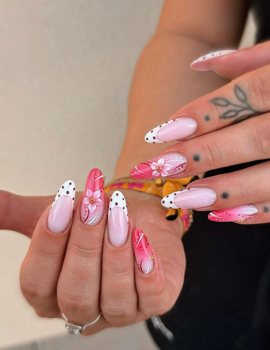Pink almond nails with black dotted white tips, bright pink floral accents, and small yellow detail