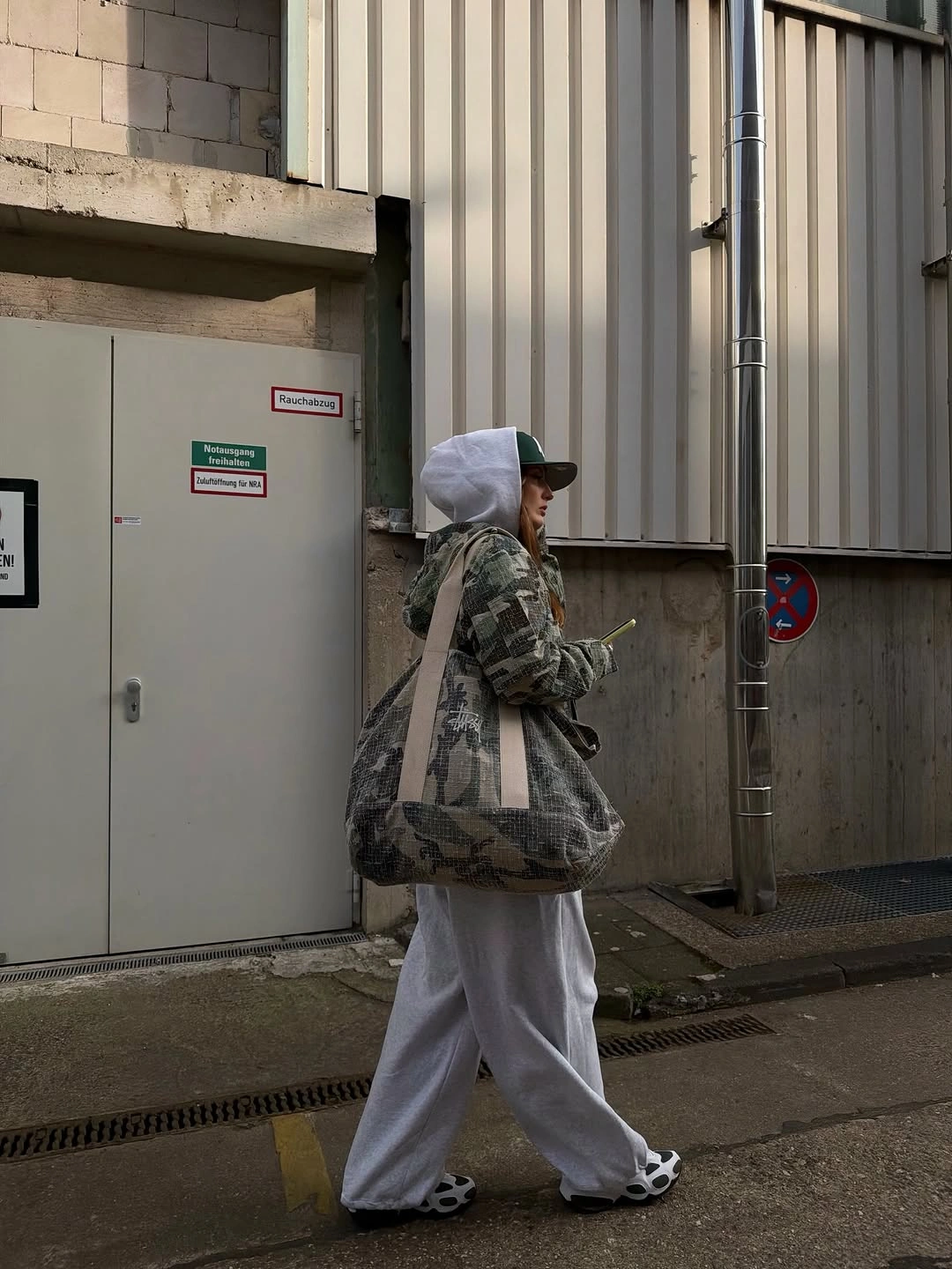 Camouflage hooded jacket with loose gray sweatpants, dark patterned tote, and dark shoes