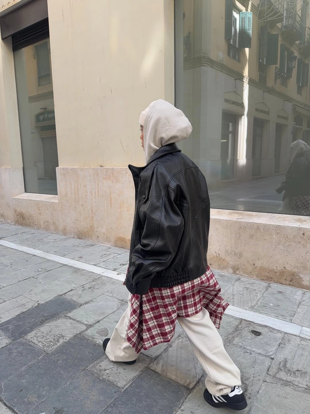 Black jacket layered over long red plaid shirt, white hood, loose pale trousers, and dark sneakers