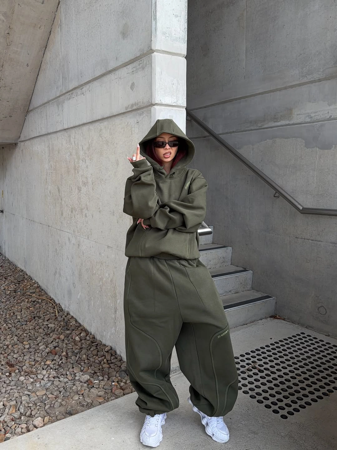 Olive vest layered over olive hoodie and matching jogger pants with white sneakers and dark sunglasses