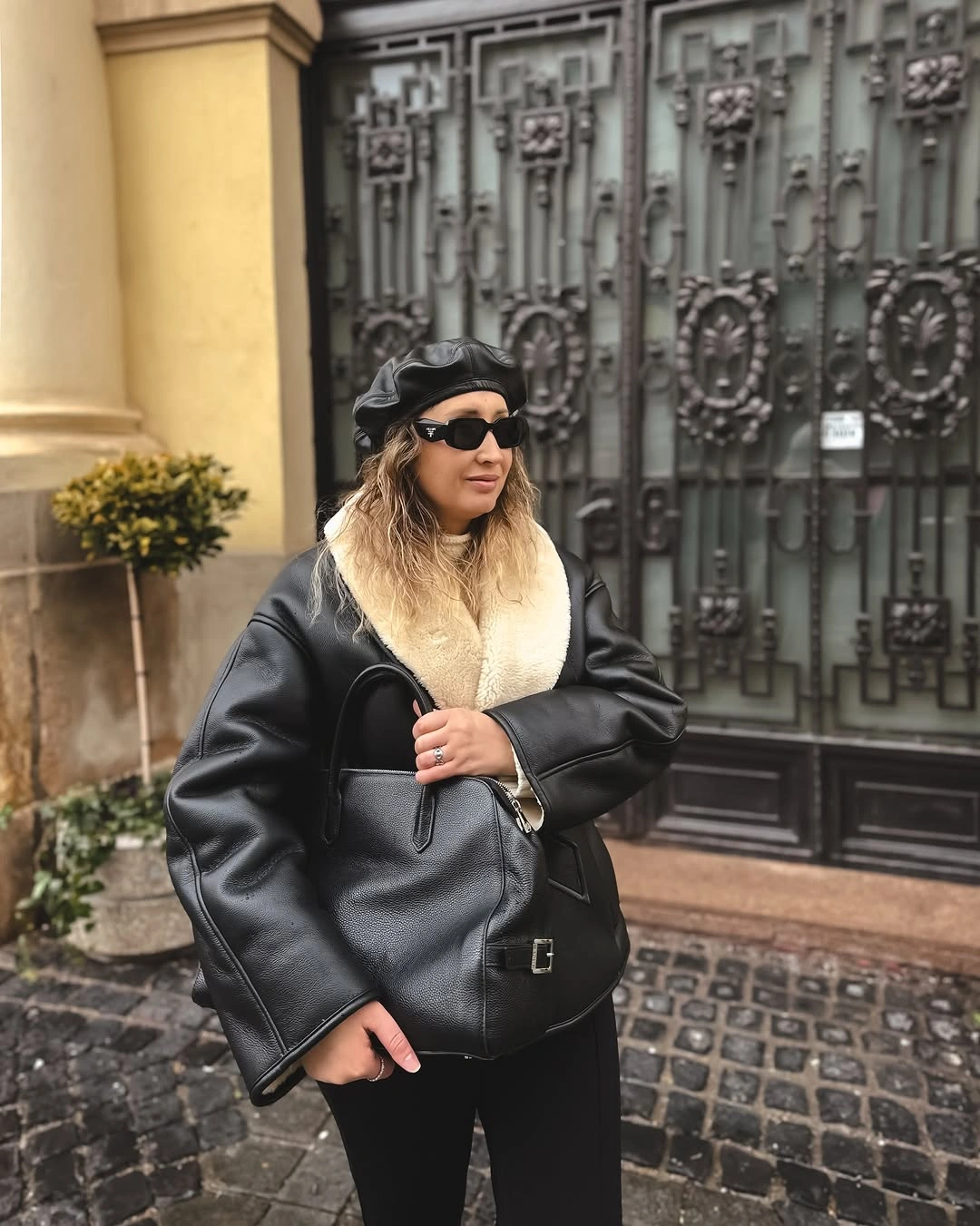 Oversized glossy black jacket with thick cream scarf, dark peaked hat, sunglasses, and compact black bag