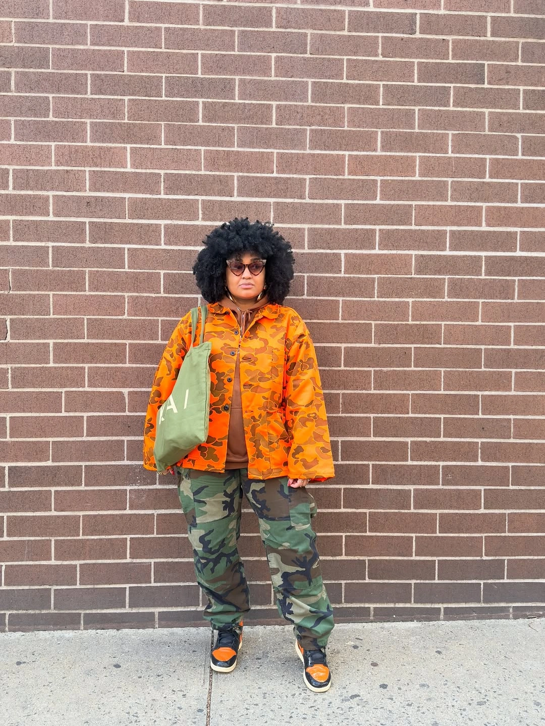 Orange printed jacket with camouflage trousers, orange black sneakers, green bag, and sunglasses