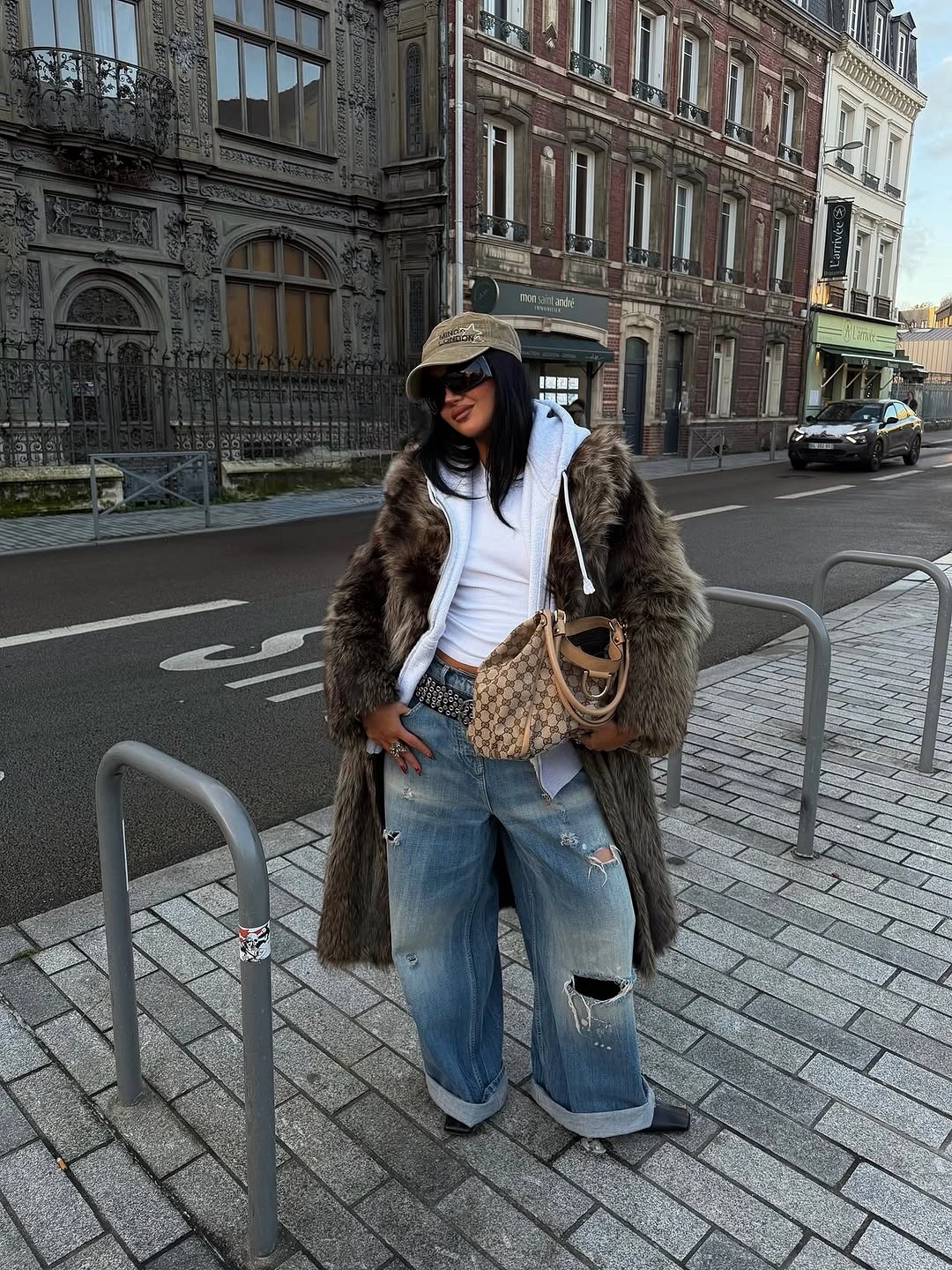 Long brown textured coat over white hoodie with distressed loose jeans, dark cap, and tan woven bag
