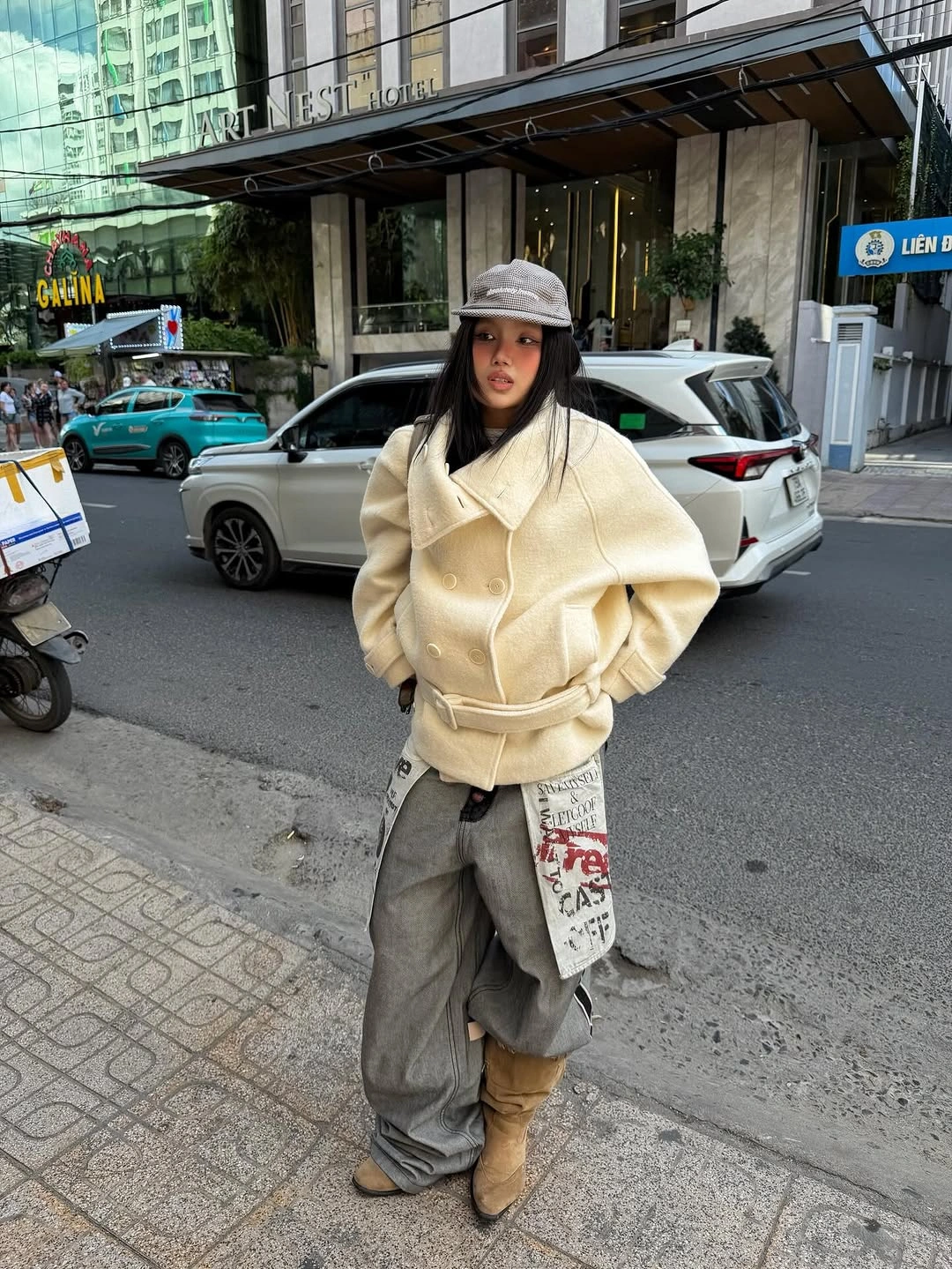 Cream belted jacket with gray cargo pants, tan boots, black cap, and patterned pouch
