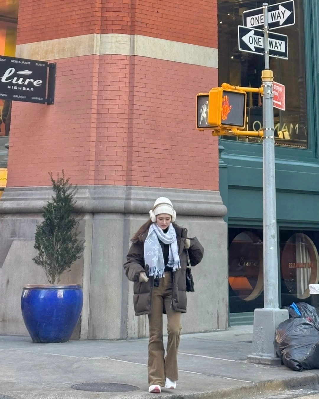 Person in brown outerwear, pale scarf and earmuffs on city corner