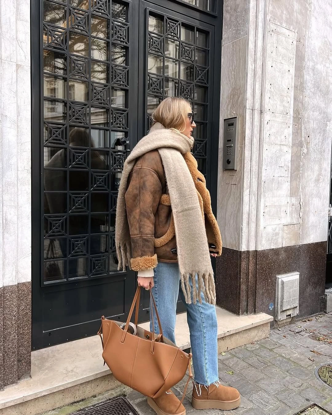 Brown trimmed jacket with cream scarf, light jeans, tan tote, and ankle boots